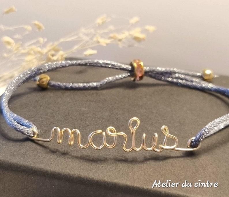 May include: A blue adjustable bracelet with a gold wire name "marlus" and a pink crystal bead.