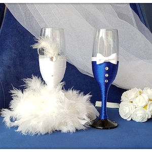 May include: Two champagne flutes decorated for a wedding. The bride's flute is wrapped in white fabric and feathers, with a blue ribbon and rhinestones. The groom's flute is wrapped in blue fabric with a white bow tie and rhinestones.