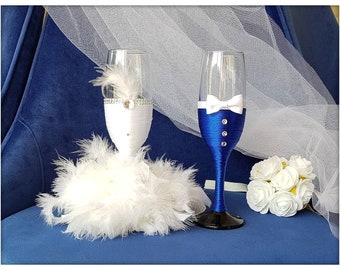 May include: Two champagne flutes decorated for a wedding. The bride's flute is wrapped in white fabric and feathers, with a blue ribbon and rhinestones. The groom's flute is wrapped in blue fabric with a white bow tie and rhinestones.