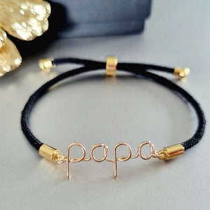Puede incluir: Una pulsera de cord&oacute;n negro con detalles dorados y la palabra "papa" en delicada escritura de alambre. La pulsera presenta extremos ajustables dorados y una cuenta deslizante para ajustar la talla. Un accesorio elegante.