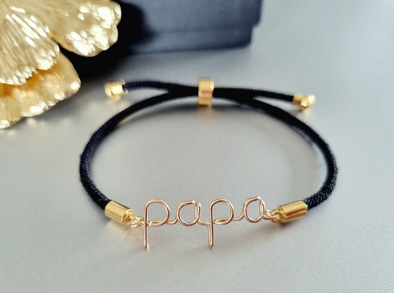 Puede incluir: Una pulsera de cord&oacute;n negro con detalles dorados y la palabra "papa" en delicada escritura de alambre. La pulsera presenta extremos ajustables dorados y una cuenta deslizante para ajustar la talla. Un accesorio elegante.