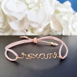 May include: A pink string bracelet with the name "Roxanne" written in gold wire.