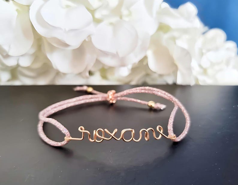 May include: A pink string bracelet with the name "Roxanne" written in gold wire.