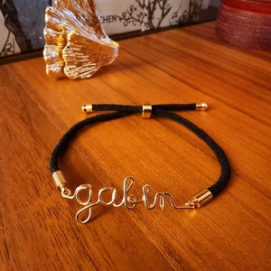 Puede incluir: Una pulsera de cord&oacute;n negro con detalles met&aacute;licos dorados y el nombre "gavin" escrito con alambre dorado. La pulsera se muestra sobre una superficie de madera.