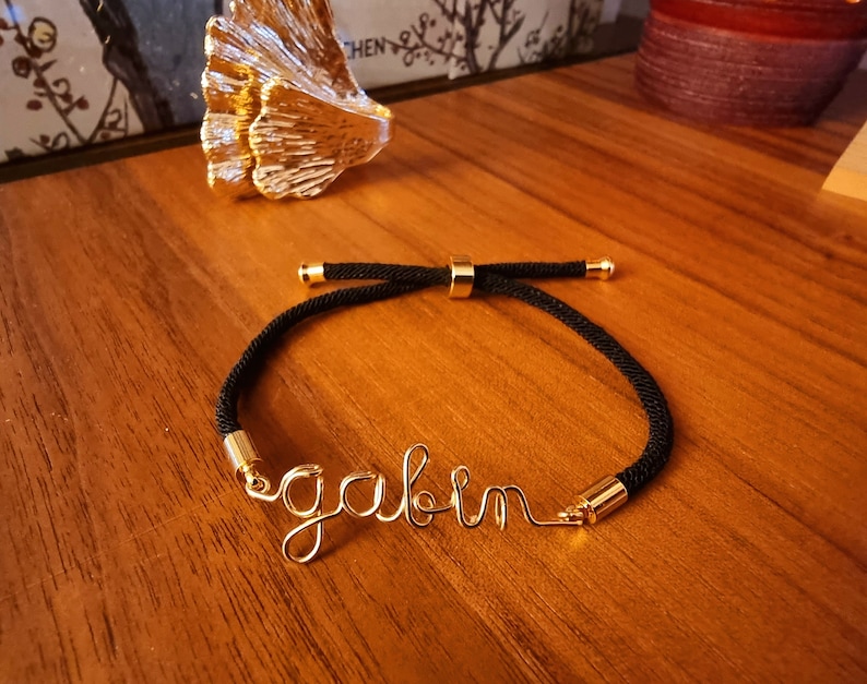Puede incluir: Una pulsera de cord&oacute;n negro con detalles met&aacute;licos dorados y el nombre "gavin" escrito con alambre dorado. La pulsera se muestra sobre una superficie de madera.
