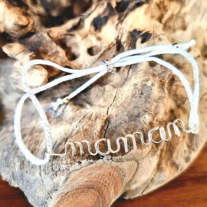 May include: White string bracelet with a silver wire word "maman" charm. The bracelet is tied around a piece of driftwood.