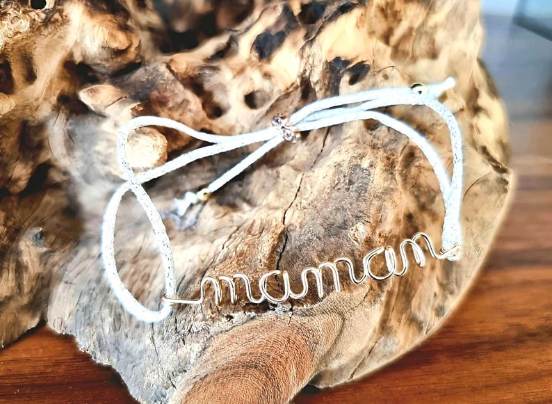May include: White string bracelet with a silver wire word "maman" charm. The bracelet is tied around a piece of driftwood.