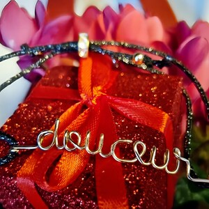 May include: A gold-colored wire bracelet with the word "delice" spelled out. The bracelet is on top of a red gift box with a red ribbon. The gift box is surrounded by pink flowers.