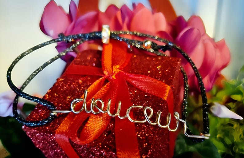 May include: A gold-colored wire bracelet with the word "delice" spelled out. The bracelet is on top of a red gift box with a red ribbon. The gift box is surrounded by pink flowers.