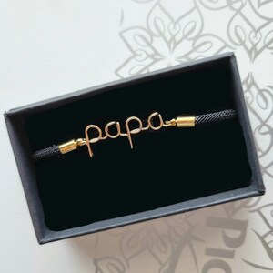 Puede incluir: Una pulsera de alambre de color dorado con la palabra "papa" en una cuerda negra. La pulsera se presenta en una caja de regalo negra. La palabra "papa" est&aacute; escrita en cursiva.