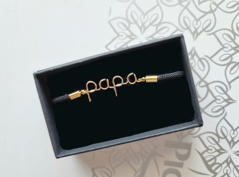 Puede incluir: Una pulsera de alambre de color dorado con la palabra "papa" en una cuerda negra. La pulsera se presenta en una caja de regalo negra. La palabra "papa" est&aacute; escrita en cursiva.