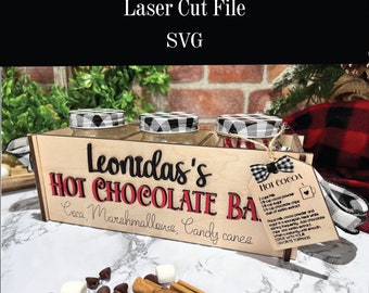 May include: A wooden box with three glass jars, labeled "Leonidas's Hot Chocolate Bar", with the text "Coca, Marshmallows, Candy canes" underneath. The jars have black and white checkered lids. A tag with the text "Hot Cocoa" and a recipe is attached to the box.