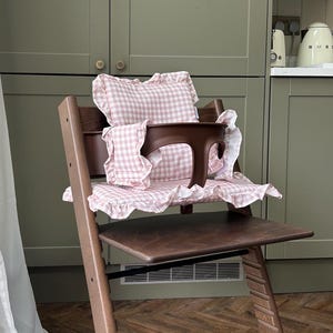 May include: A dark brown wooden high chair featuring a removable dark brown plastic safety bar and a soft, light pink and white gingham fabric cushion set with ruffled edges, including a backrest pillow and a seat pad for added comfort.