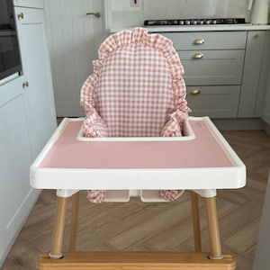 May include: A baby high chair with a pink tray and a gingham patterned seat. The chair has light wood legs and a white frame. The seat has a ruffled edge. The high chair is in a kitchen setting.