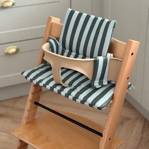 Pode incluir: Uma cadeira alta de madeira com assento bege e almofada listrada em tons de verde e branco. A cadeira alta tem apoio para os pés e barra de segurança. O fundo mostra uma cozinha de cor clara.