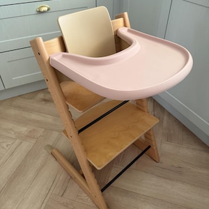 Puede incluir: Una trona de madera con una bandeja rosa claro y un respaldo beige. La silla tiene dos estantes de madera y soportes de metal negro. La silla está diseñada para un bebé o un niño pequeño.