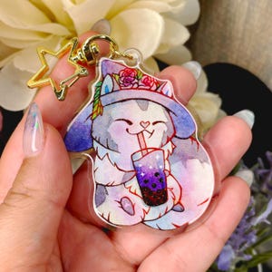 Può includere: Un portachiavi in acrilico trasparente con un gatto dei cartoni animati che indossa un cappello da strega viola e blu con fiori, che tiene un boba tea a tema galassia con una cannuccia.
