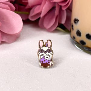 Puede incluir: Un pin de esmalte dorado con un mapache de dibujos animados que lleva un sombrero de paja y sostiene una taza de té de boba. El té de boba es morado con perlas de boba blancas y una pajita.