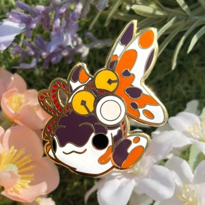 Puede incluir: Un pin de esmalte dorado con un pez de dibujos animados con detalles naranja, blanco y morado. El pez tiene ojos grandes y redondos y una expresión caprichosa.