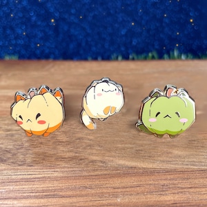Puede incluir: Tres pines de esmalte con personajes de dibujos animados. El primer pin es una calabaza amarilla con una cara linda. El segundo pin es un gato blanco con una cara linda. El tercer pin es una calabaza verde con una cara linda.