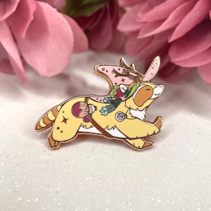 Puede incluir: Un pin de esmalte dorado con un animal amarillo que lleva una corona de flores rosa y blanca, llevando una pequeña bolsa marrón. El animal tiene una nariz rosa y ojos marrones. El pin es un diseño caprichoso y colorido.