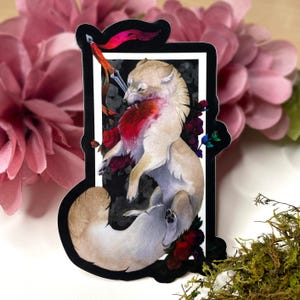 Puede incluir: Una pegatina con una ilustración en acuarela de un lobo atravesado por una espada. El lobo está representado en tonos beige y blanco, con un toque de rojo. Rosas rojas oscuras y un marco negro rodean al lobo.