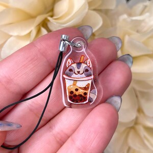 May include: A clear plastic charm with a cartoon cat inside a boba tea cup. The cat is gray and white with brown eyes. The boba tea is brown with white boba pearls. The charm has a black string attached to it.