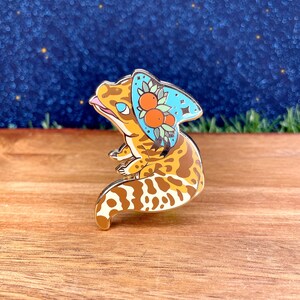 Può includere: Uno spillo in smalto dorato con un geco dei cartoni animati che indossa un cappello blu e bianco con fiori arancioni. Il geco ha un motivo a macchie marroni e bianche.