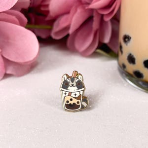 Puede incluir: Pin de esmalte dorado con un mapache de dibujos animados que lleva un sombrero marrón y blanco, dentro de una taza transparente con un líquido marrón y perlas de boba. El mapache está mirando hacia arriba con una expresión feliz.