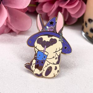 May include: A gold-tone enamel pin featuring a cartoon animal wearing a purple and blue witch hat with stars and flowers. The animal is holding a blue and purple boba tea with a straw.