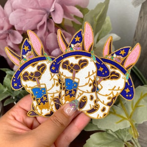 Puede incluir: Tres parches bordados para planchar que representan un conejo blanco con un sombrero de mago azul y morado y que sostiene una copa azul y dorada. El conejo tiene ojos negros y una nariz rosa.