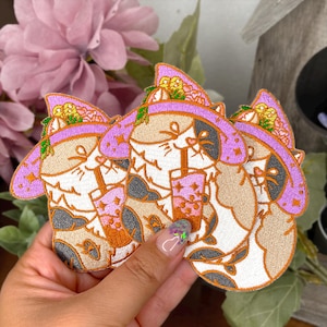 Puede incluir: Tres parches bordados para planchar que representan un gato de dibujos animados con un sombrero de bruja morado y sosteniendo un té de boba rosa y blanco. El gato tiene orejas negras y una cara blanca con pelaje naranja.