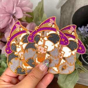 Może przedstawiać: Trzy haftowane łatki z szopami w fioletowych kapeluszach czarodzieja pijących boba tea. Łatki są wykonane z filcu i mają szczegółowy wzór.