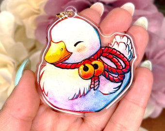 May include: A white duck with a red rope and two gold bells around its neck. The duck is drawn in a cartoon style with a sparkly finish. The duck is a charm on a clear acrylic keychain.