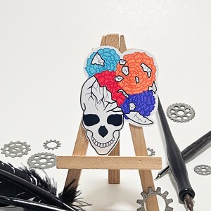 May include: A white skull sticker with a colorful floral design on top. The sticker is on a small wooden easel. The background is white with black feathers and metal gears.