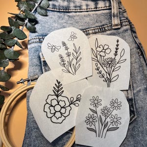 May include: Four embroidery patterns on white paper, featuring floral designs with bees, placed on a pair of blue denim jeans.