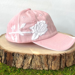 May include: A pink baseball cap with a white embroidered rose and stem design. The cap has a curved brim and a white underside.
