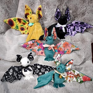 Peut inclure: Cinq chauves-souris en peluche avec des corps doux et des ailes à motifs. L'une est jaune avec des tournesols, une noire avec des motifs d'Halloween violets, une sarcelle avec des fleurs roses, une blanche avec des symboles occultes noirs.
