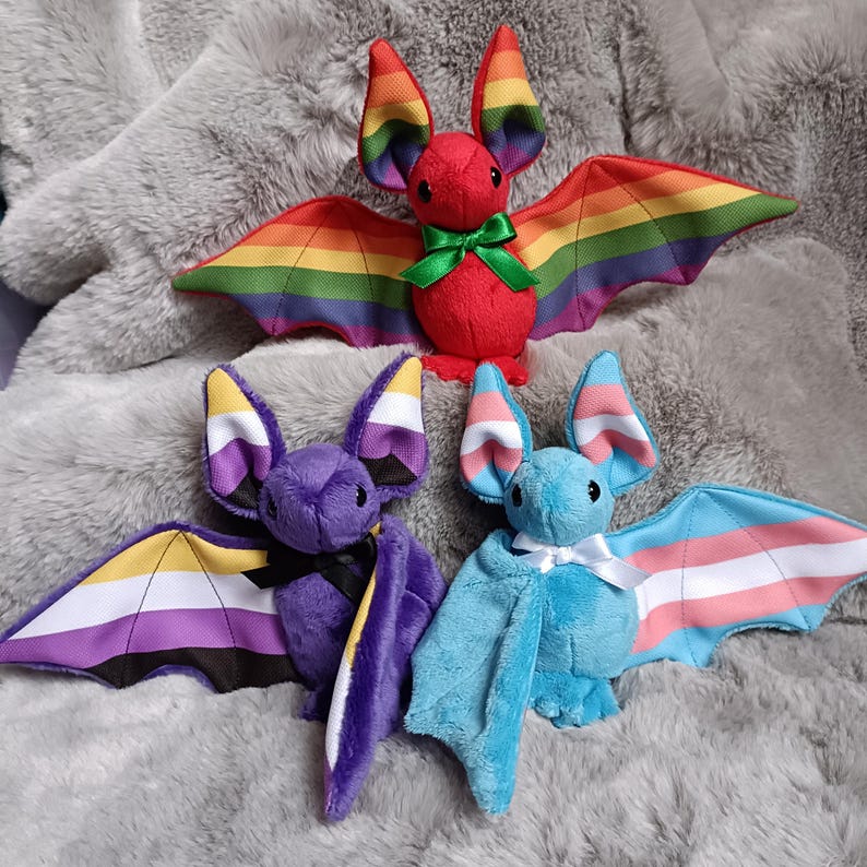 Puede incluir: Tres murci&eacute;lagos de peluche con cuerpos de tela suave. El murci&eacute;lago superior es rojo con alas arco&iacute;ris y un lazo verde. El murci&eacute;lago inferior izquierdo es morado con alas de bandera no binaria y un lazo negro. El murci&eacute;lago inferior derecho es azul con alas de bandera trans y un lazo blanco.