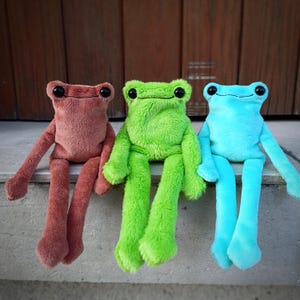 Puede incluir: Tres juguetes de peluche con forma de rana en tonos rojo-marrón, verde y azul. Cada rana tiene grandes ojos negros y extremidades largas y colgantes. Las ranas están sentadas una al lado de la otra, con las piernas colgando. Los peluches son de material suave y difuso.