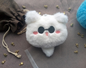May include: A white plush bear-shaped keychain with black sunglasses, pink cheeks, and a small tail. The keychain is surrounded by small gold star charms and a brown drawstring bag. A blue pom-pom is visible in the background.