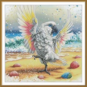 May include: A cross-stitch artwork depicts a white cockatoo with outstretched wings, showcasing yellow, pink, and red feathers. The bird stands on a sandy beach with seashells, the ocean in the background. The artwork is framed in a gold-colored border.