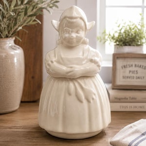 May include: Cream-colored ceramic cookie jar shaped like a Dutch girl, with arms crossed. The jar has a lid and is on a wooden surface. The jar is in a kitchen setting with a sign that says "FRESH BAKED PIES SERVED DAILY".