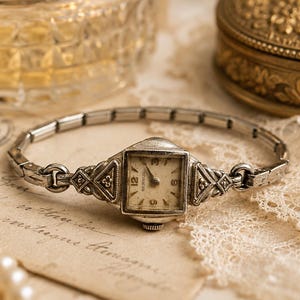May include: A silver-toned Benrus wristwatch with a square face, featuring a cream-colored dial with black numerals and hands. The watch has an intricate, geometric metal link band and decorative triangular lugs connecting the case to the strap.