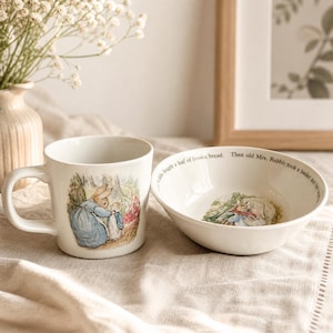 May include: White ceramic mug and bowl set featuring illustrations of rabbits in a garden scene. The mug has a handle and the bowl has text around the rim. The set is on a neutral-colored surface.