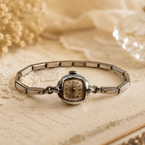 May include: A silver-toned analog wristwatch with a square-shaped face, featuring a light-colored dial with black numerals and a metal link bracelet. The watch has a small winding crown at the top and a polished finish on the case and band.