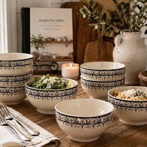 May include: A set of cream-colored ceramic bowls featuring a black decorative filigree pattern around the exterior rim. The collection includes several stacked bowls and individual bowls, some containing food, arranged on a wooden surface.