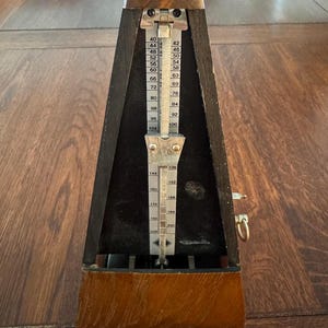 May include: A pyramid-shaped wooden metronome with a dark brown finish. It features a central vertical metal scale with printed tempo markings ranging from 40 to 208 beats per minute, a sliding metal weight, and a small gold-toned metal ring on the side.