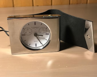 May include: A silver-toned Markman quartz travel clock with a black leather case. The clock face is white with black numerals and hands, and features a second dial and an alarm indicator. The clock is rectangular and has a loop for hanging.