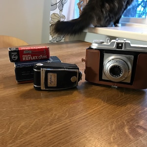 May include: A collection of vintage cameras and film boxes on a wooden surface. The cameras include a black and silver Agfa camera and a brown leather-cased camera. Film boxes include a red and black Philips Reflex film box.